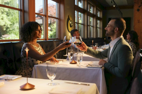A couple is toasting with wine glasses at a restaurant table. They are sitting by large windows, with another couple in the background.