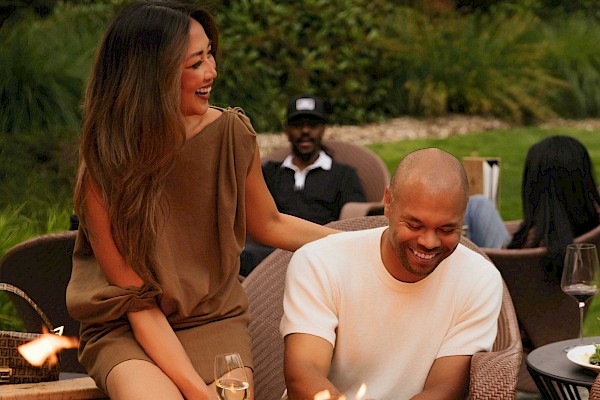 A group of people enjoying food and drinks around a fire pit outside, with two smiling in the foreground.
