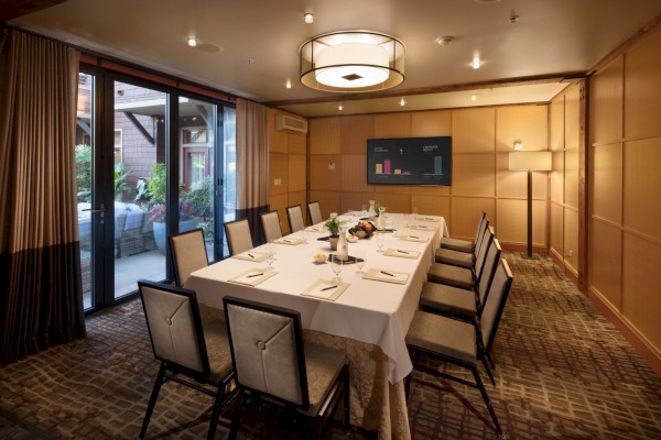A conference room with a long table, chairs, a large window, a wall-mounted screen, and ambient lighting.
