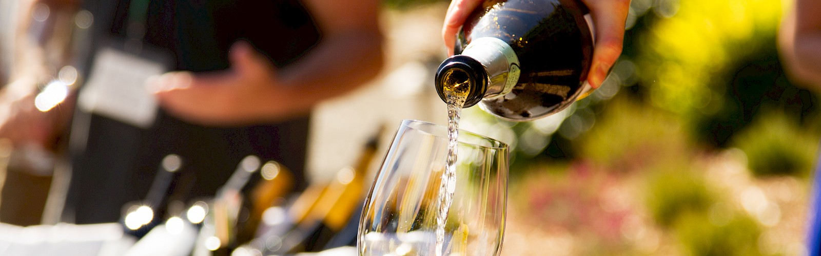 A person is pouring white wine into a glass held by another person at an outdoor event with blurred people and greenery in the background.