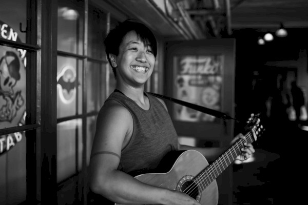 A person smiling while playing an acoustic guitar in a dimly lit room with posters on the walls.