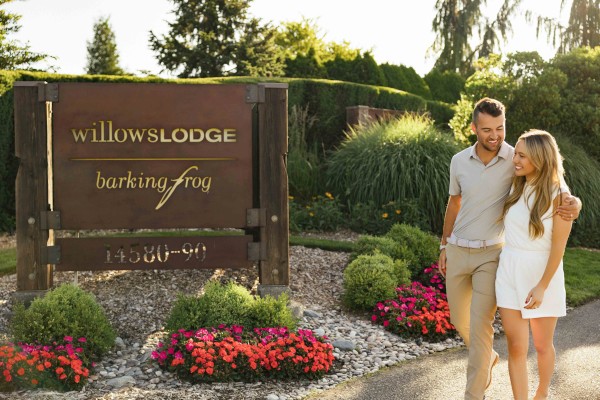 A couple walking near a sign for Willow Lodge and Barking Frog, surrounded by lush greenery and flowers.