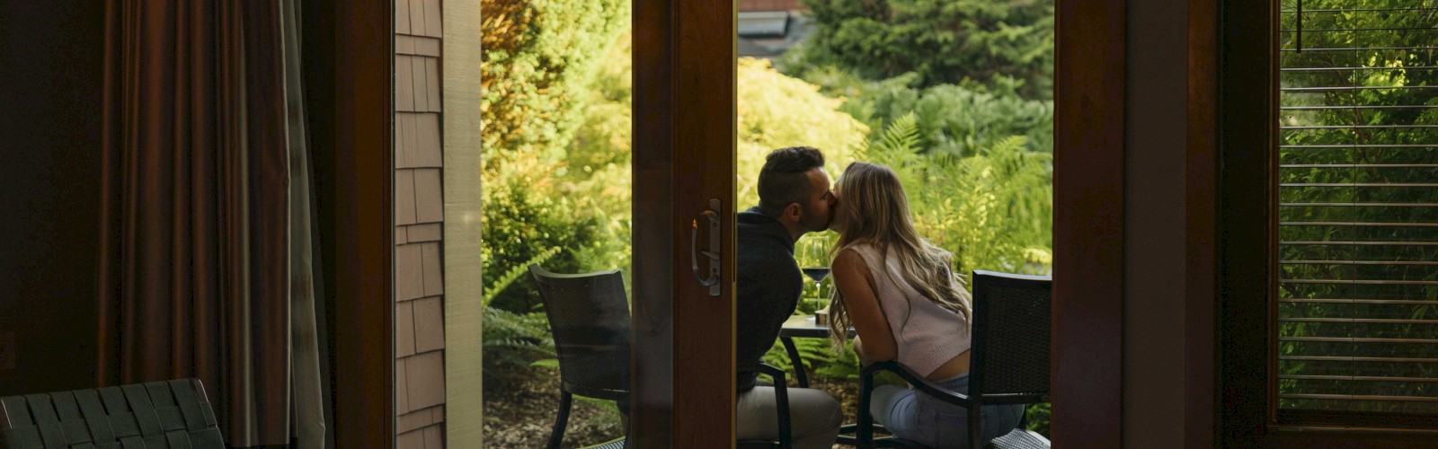 A couple is sitting outside on a patio, leaning in for a kiss, with a garden view from inside a room through open doors.