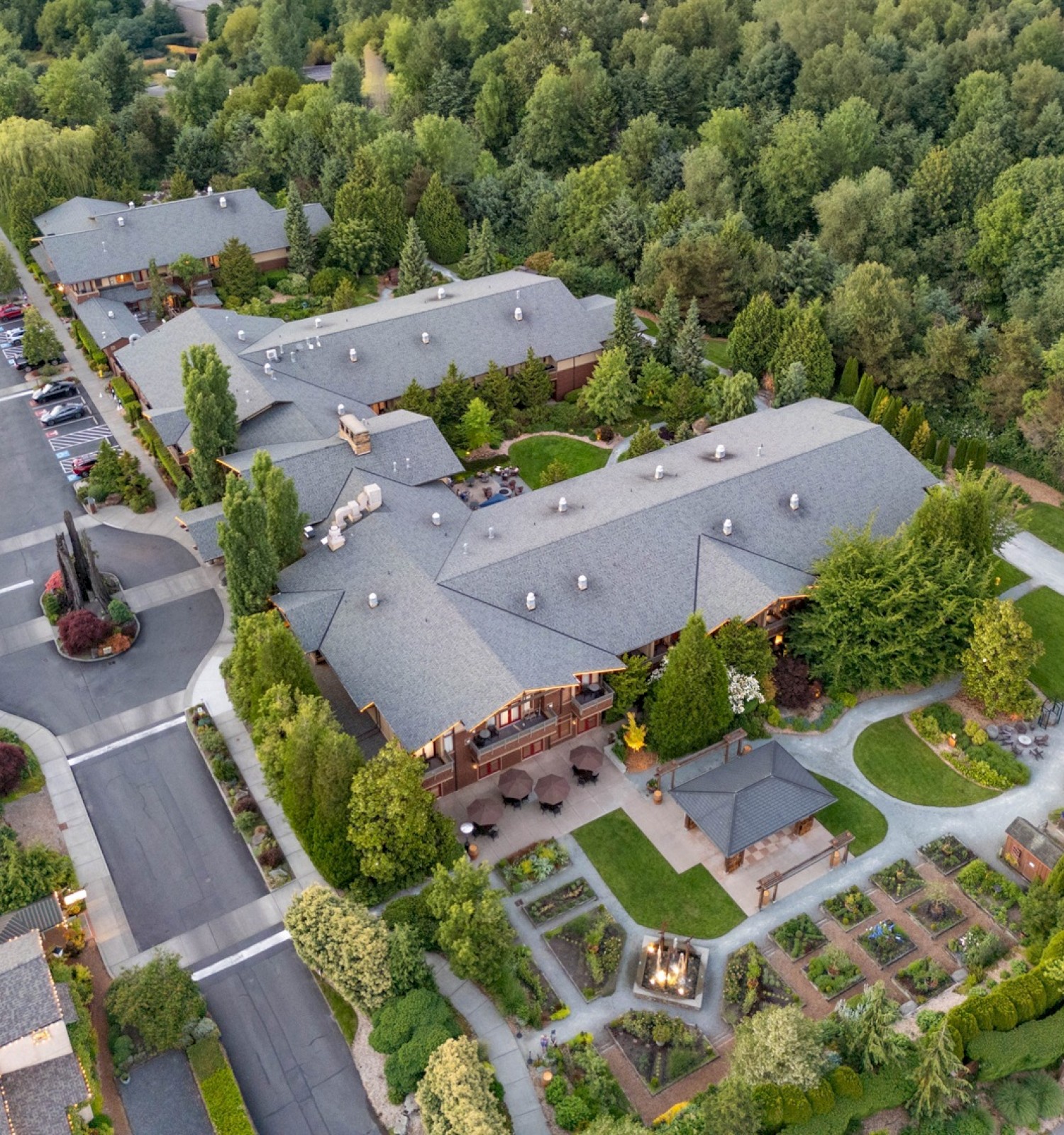 Aerial view of a building complex surrounded by trees, parking lots, and gardens. Paths and greenery create a tranquil setting.