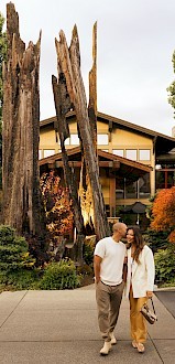 A couple stands together in front of a house with large wooden sculptures and lush greenery in the background.