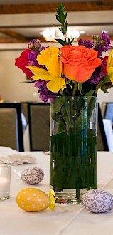 A floral arrangement in a tall glass vase with colorful flowers, decorated eggs, and a candle on a table, likely for a celebration or holiday.