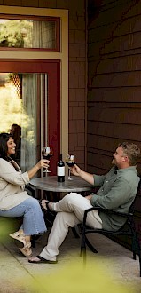 A couple enjoys a sunny outdoor date on a porch, clinking wine glasses at a small table with a bottle between them, cozy and relaxed.