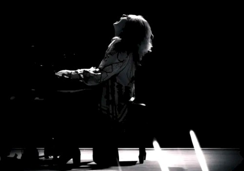 A person seated at a piano on a stage, silhouetted dramatically, with streaks of light across the foreground creating a dynamic effect.