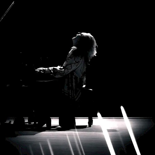 A person seated at a piano on a stage, silhouetted dramatically, with streaks of light across the foreground creating a dynamic effect.