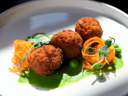 This image shows three crispy fried balls served with shredded carrots, green peas, and a green sauce on a white plate.