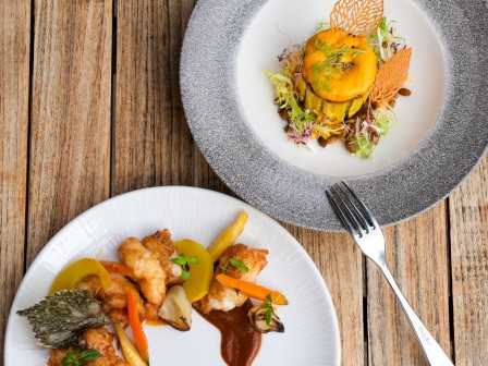The image shows two plates of gourmet food on a wooden table, featuring colorful, artistically presented dishes with garnishes and sauces.