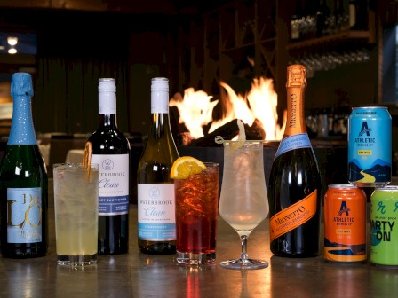 A row of assorted drinks: sparkling wine, cocktails, and bottled mixers on a bar counter with a flame-lit background, ending with a bright orange can.
