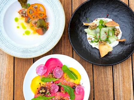 The image shows three colorful, artistically plated dishes of gourmet food on a wooden table.