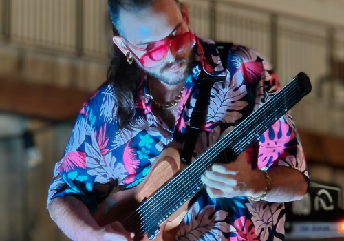 A person in a floral shirt is playing an unusual string instrument, possibly a multi-string guitar, outdoors with railing in the background.