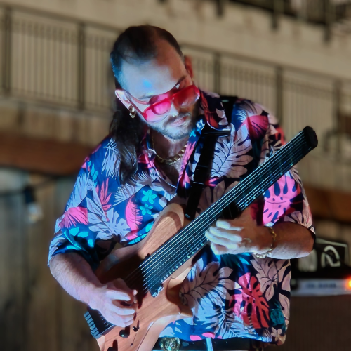 A person in a floral shirt is playing an unusual string instrument, possibly a multi-string guitar, outdoors with railing in the background.