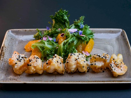 A dish with tempura shrimp garnished with sesame seeds and greens, accompanied by oranges on a rectangular plate.
