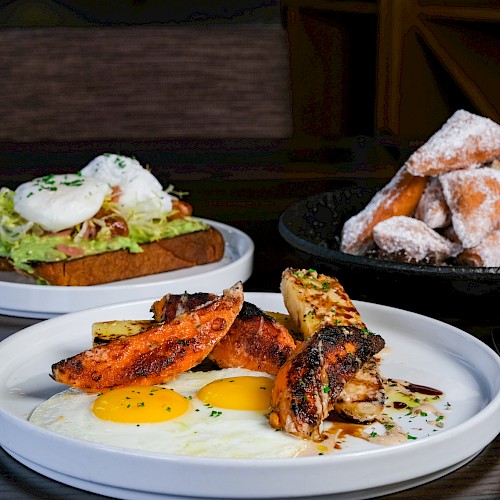 A breakfast plate with eggs, grilled chicken, and roasted vegetables, a toast with avocado and poached eggs, and a bowl of fried pastry dusted with powdered sugar.
