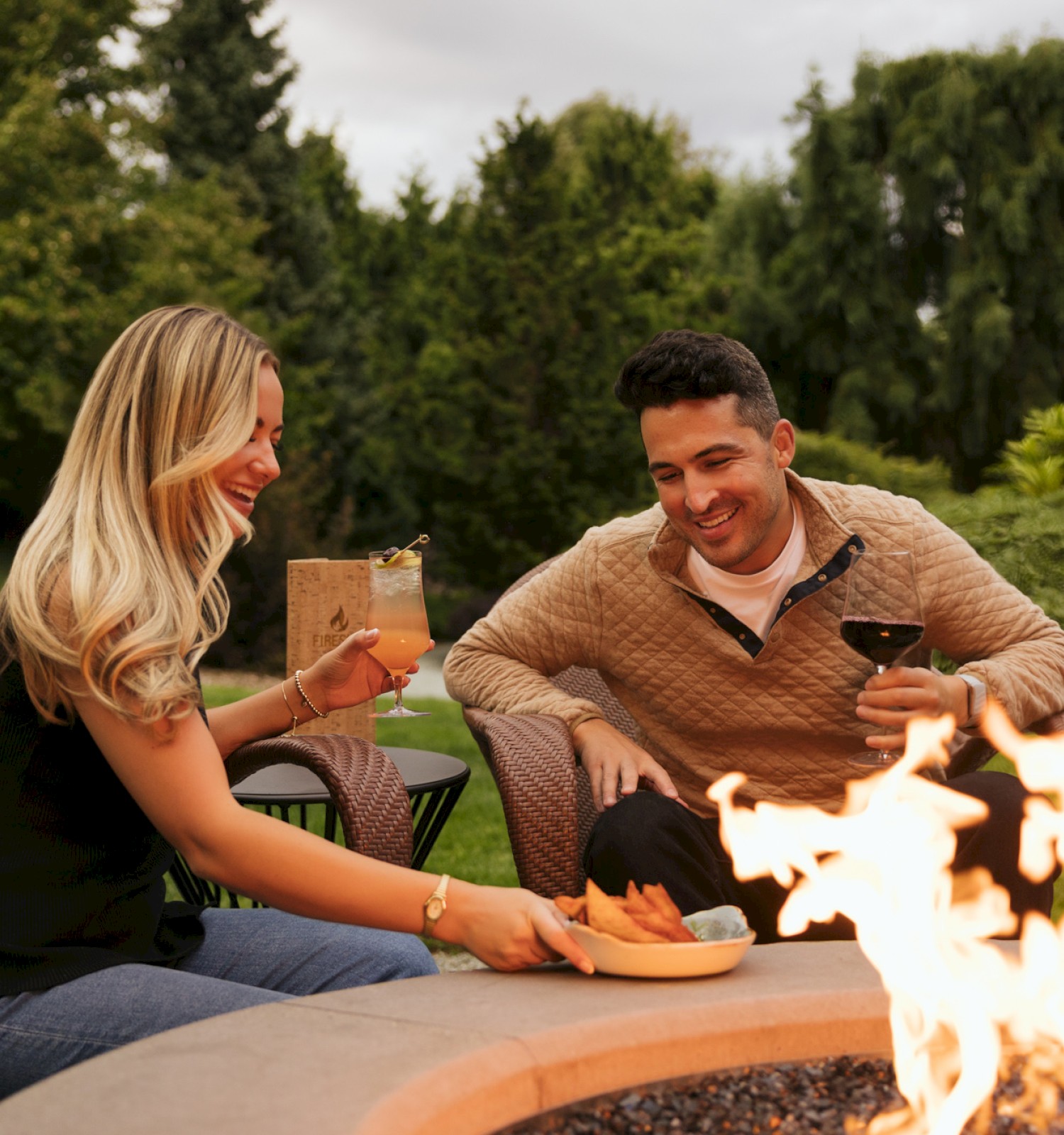 Two friends enjoy drinks, food, and laughter around a backyard fire pit on a cozy evening with lush greenery in the background.