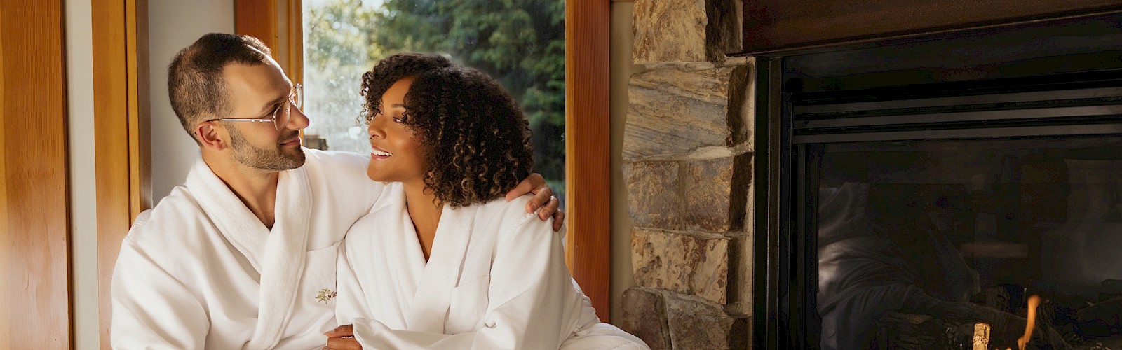 A couple in white robes sits by a fireplace with breakfast, champagne, and orange juice, looking at each other.