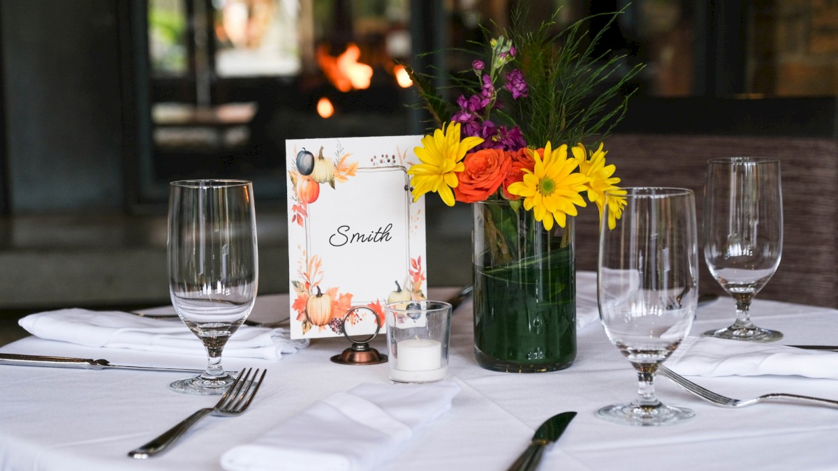 A table set for a meal with a floral centerpiece, wine glasses, utensils, a personalized place card, and a candle at a cozy event.