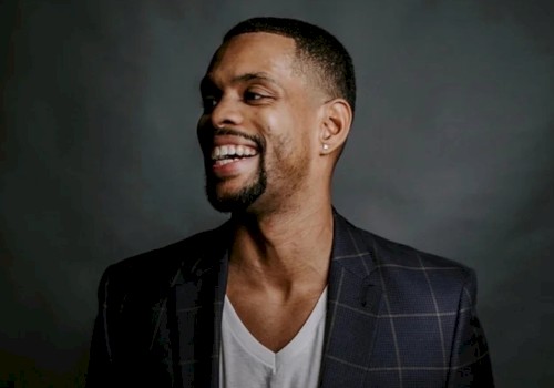 A person in a checkered suit jacket and white shirt is smiling against a dark background.