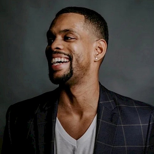 A person in a checkered suit jacket and white shirt is smiling against a dark background.