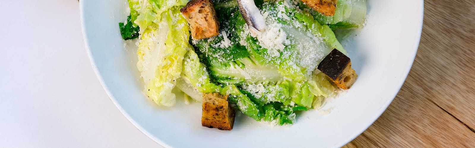 A Caesar salad with romaine lettuce, croutons, anchovies, Parmesan, and a lemon wedge in a white bowl on a wooden surface.