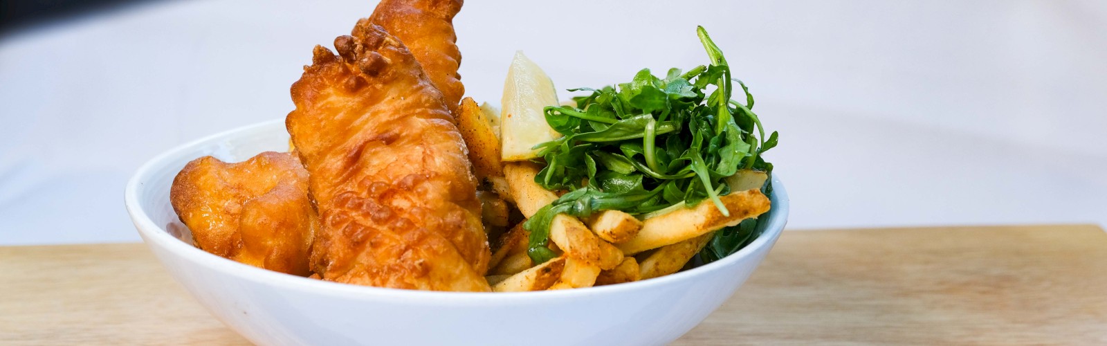 A bowl with fried fish, french fries, greens, and a lemon wedge sits on a wooden board.
