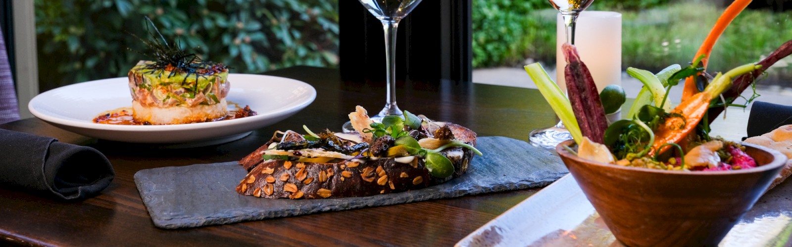The image shows a table with gourmet dishes, two glasses of white and rosé wine, and a garden view through large windows.