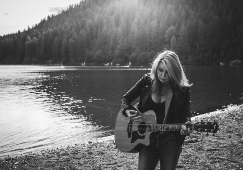 A woman stands on a lakeshore playing an acoustic guitar, with trees and hills in the background and sunlight streaming over the water.
