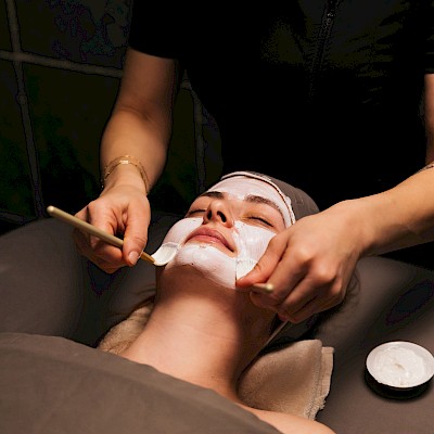 A person lies back with a facial mask being applied by a technician using brushes and a headband, while resting on a padded spa bed.