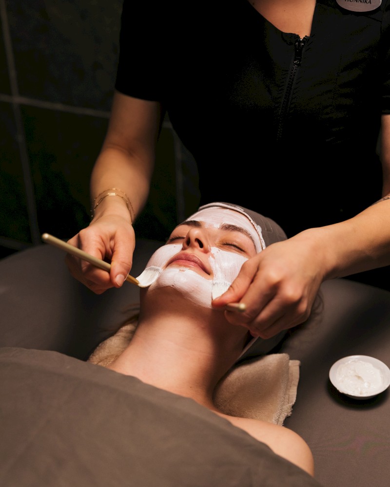A person lies with a white facial mask while a practitioner applies product and tools, performing a spa facial treatment.