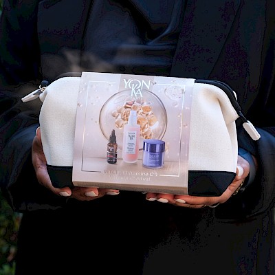 A person in dark clothing holding a small white cosmetic bag with a label and bottle images on the front, outdoors.