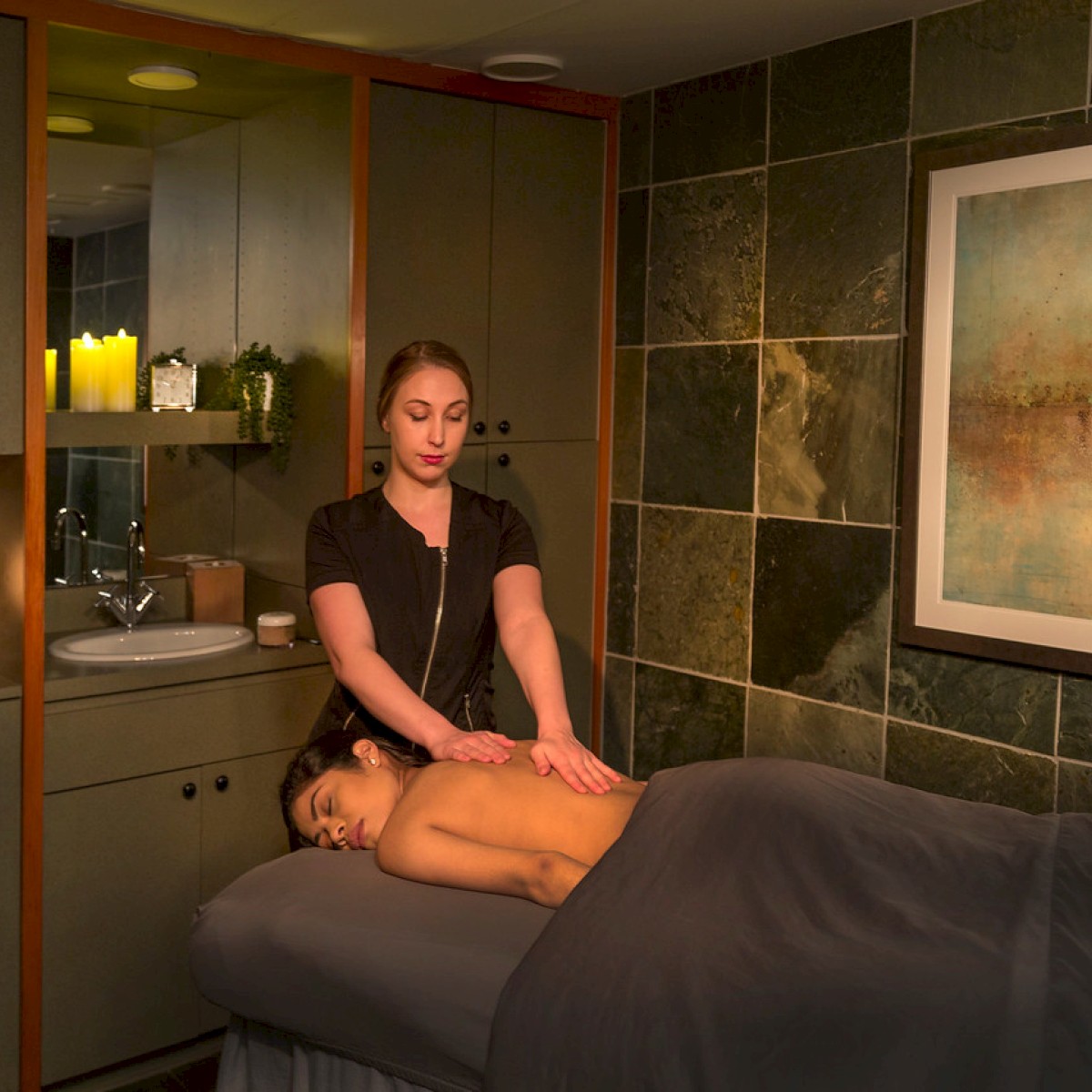 A woman receives a massage in a relaxing spa room with candles, paintings, and a sink in the background.