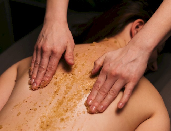 Two hands massage a back with a brown exfoliating scrub, in a spa setting, while the person lies face down, relaxing.