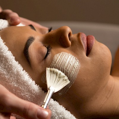 A woman receives a facial treatment, with a brush applying a skincare product onto her cheek for relaxation or skincare purposes.