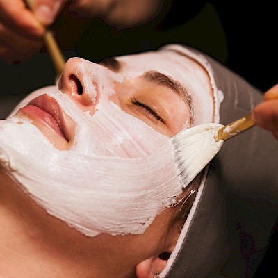 A person receiving a facial treatment: a white facial mask is being applied with brushes on the face, eyes closed.