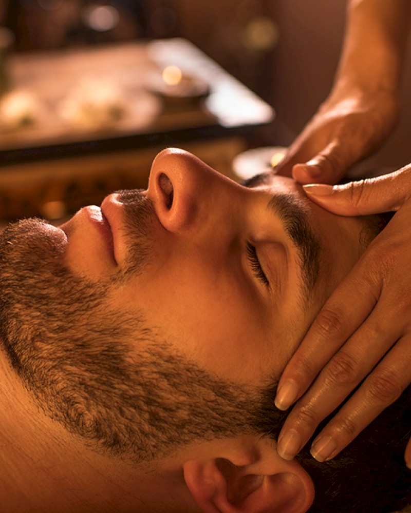 A man receives a relaxing massage on his face in a cozy, candle-lit spa setting.