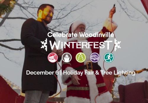 A Winterfest event banner with two people on stage, one in a suit and the other in a festive outfit, promoting Celebrate Woodinville Winterfest on December 6 with holiday fair and tree lighting.