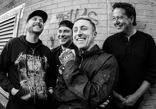 Four men stand by a brick wall, chatting and smiling, one wearing a hoodie with a graphic and another in a jacket, casual scene.