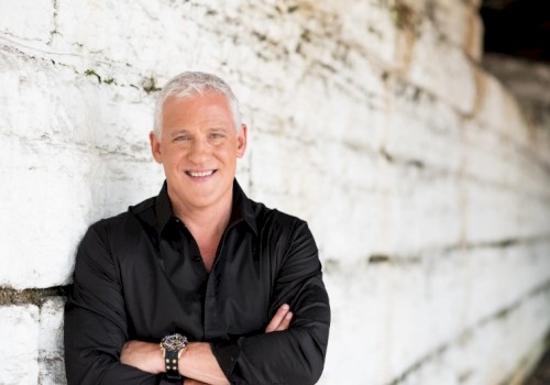A middle-aged man with short gray hair, wearing a black shirt, arms crossed, standing against a white textured wall, smiling.