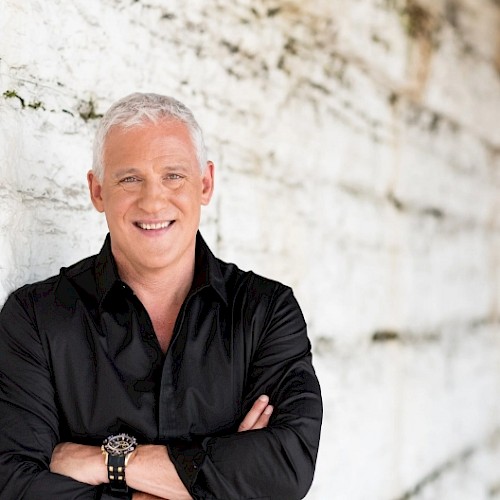 A middle-aged man with short gray hair, wearing a black shirt, arms crossed, standing against a white textured wall, smiling.