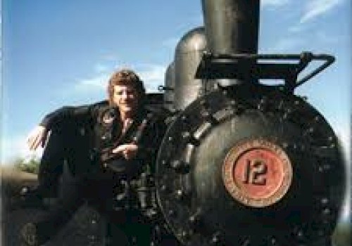 A man sits beside a vintage steam locomotive with a large black smokestack and red number plate, outdoors under a blue sky.