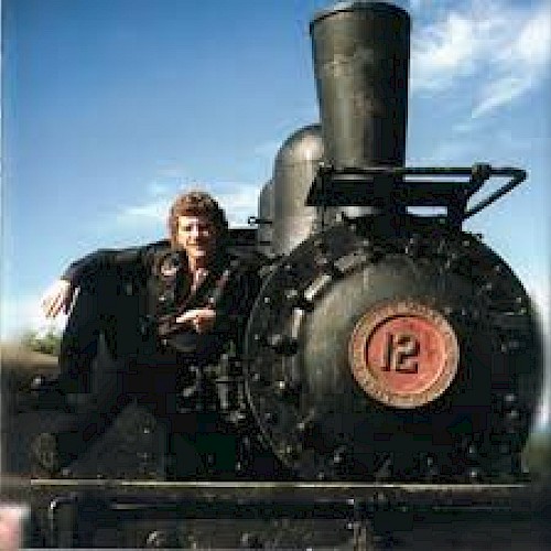 A man sits beside a vintage steam locomotive with a large black smokestack and red number plate, outdoors under a blue sky.