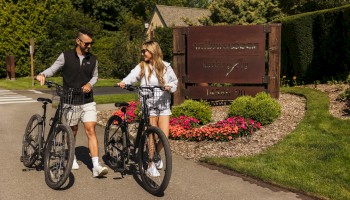 Two people ride electric bikes along a driveway past a wooden sign and well‑kept landscaping, smiling and chatting as they pedal together.