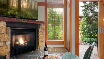 Cozy living room with a stone fireplace and mounted TV, a small table with wine, cheese, and crackers, and a glass door opening to a sunny outdoor garden.