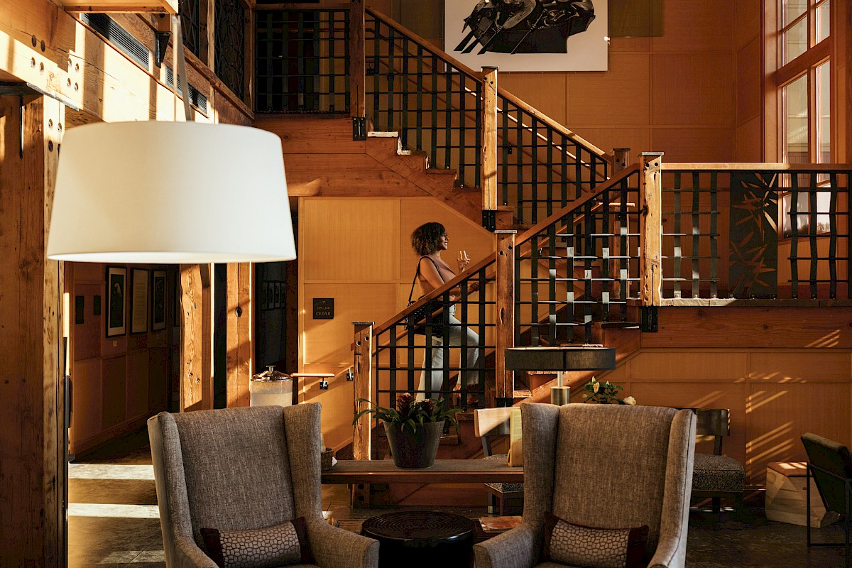 A stylish living area with warm sunlight, two gray armchairs, a tall white lamp, and a wooden staircase in a cozy, sunlit home.