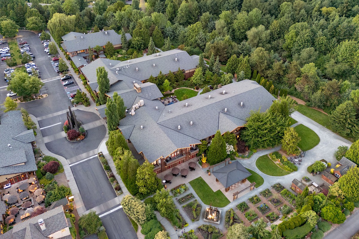Aerial view of a suburban apartment complex with gray rooftops, landscaped courtyards, and surrounding green trees; winding roads and parking lots nearby.
