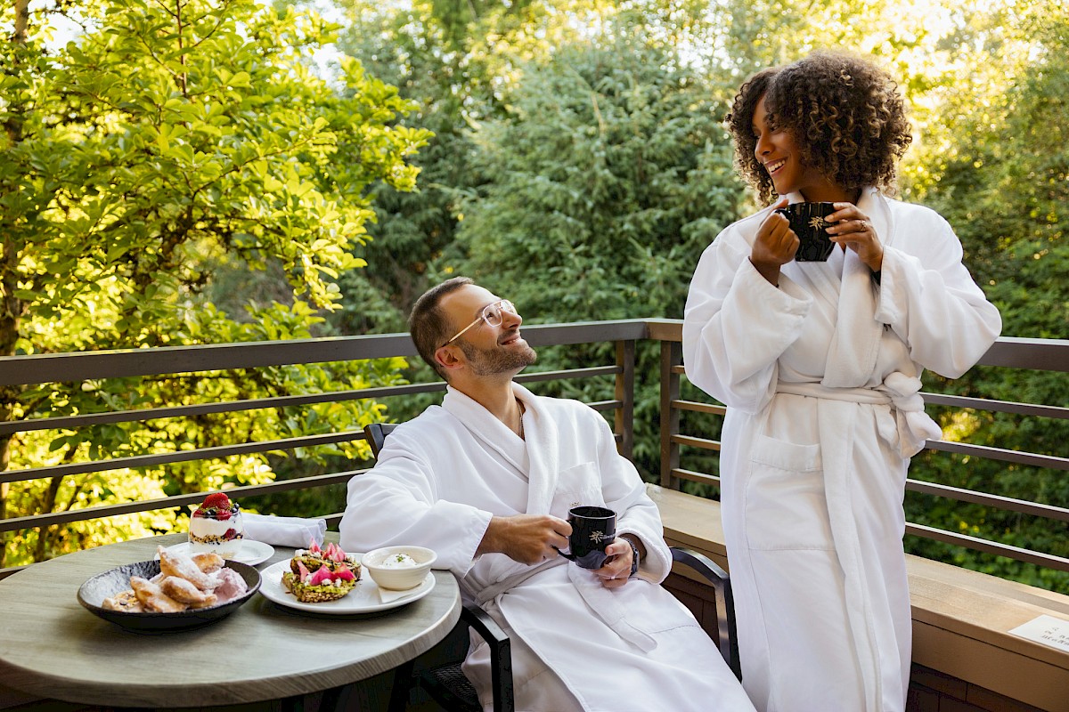 Two people in white robes enjoy coffee and pastries on a balcony deck, chatting and smiling while flowers and trees glow in the background.