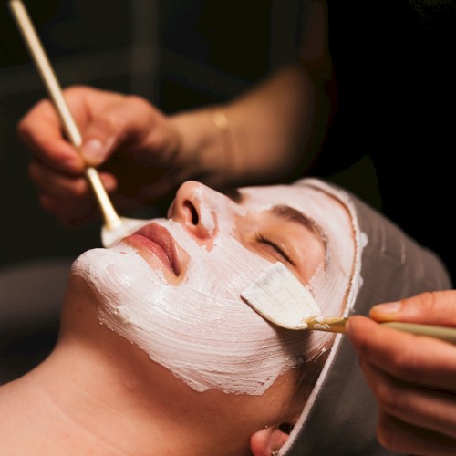 A woman is receiving a facial treatment, with her face being painted white and her eyes closed.